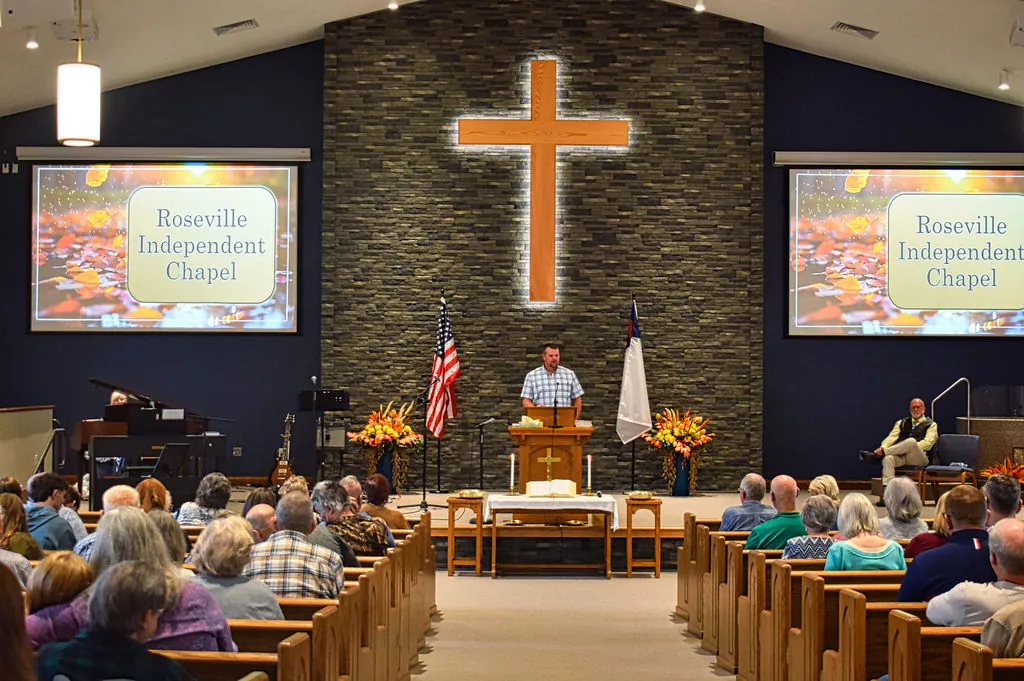 Sunday morning preaching at Roseville Independent Chapel, the lit wooden cross on the stone wall behind the pulpit